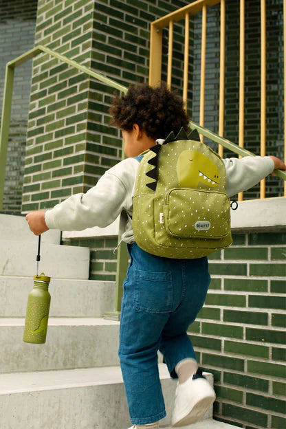 A child wearing a light green dinosaur Trixie backpack, blue jeans, and a white sweatshirt walks up outdoor concrete stairs while holding a matching light green bottle.