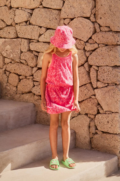 Young girl in a pink Molo dress and hat standing against a stone wall.