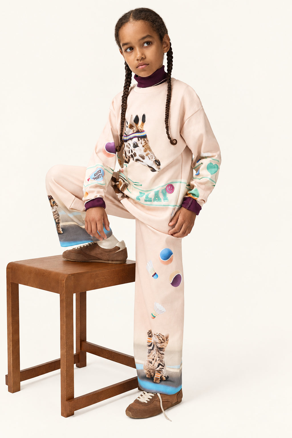 Young girl wearing a Molo pink set with animal prints sitting on a wooden stool against a white background