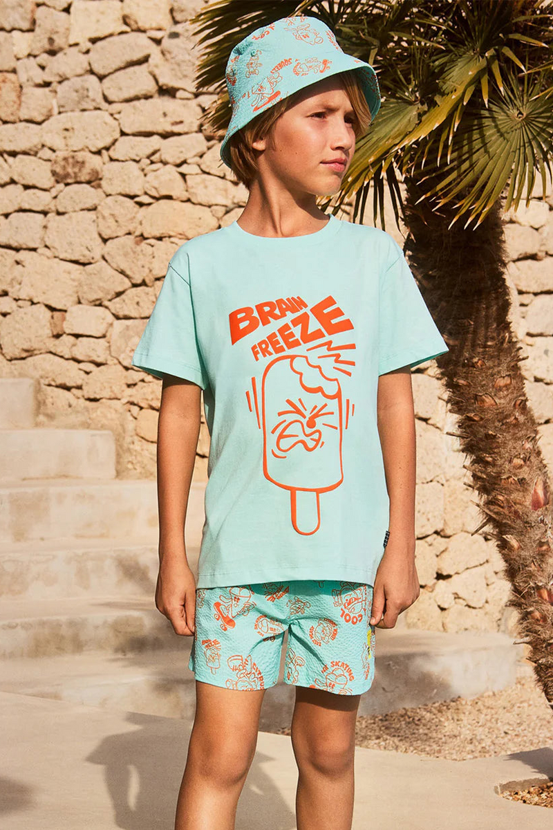Boy wearing a light blue shirt with 'Brain Freeze' design from Molo kidswear with matching shorts, standing against a stone wall.