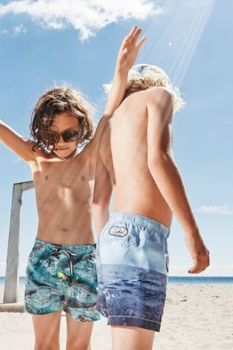 Two boys on a beach wearing patterned Molo swim shorts with a clear blue sky.